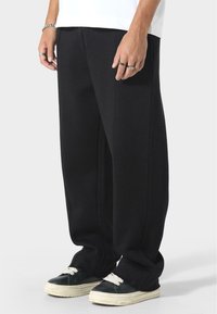 Czarne, szerokie spodnie wykonane z miękkiego materiału, z gładką strukturą i detalami w postaci fałd, w połączeniu z czarnymi trampkami i białymi sznurowadłami.