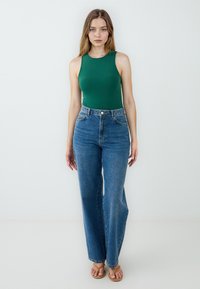 Top verde senza maniche abbinato a jeans in denim blu chiaro a vita alta. I jeans hanno una forma dritta e un leggero sbiadimento. Sandali piatti completano il look.