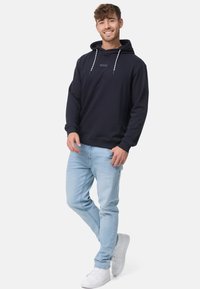 Marineblauwe hoodie met een trekkoord, kangoeroezak en textuurstof, gecombineerd met lichtblauwe jeans en witte sneakers.