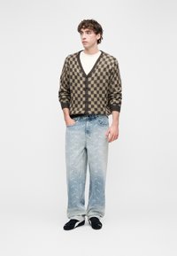 Bruin en beige geruit vest over een wit T-shirt; vervaagde blauwe spijkerbroek met lichte patronen; zwarte sneakers; neutrale achtergrond.