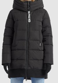 Chaqueta de plumas acolchada negra con alto cuello, cremallera frontal, bolsillos laterales y etiqueta "knu:lo" en el tirador de la cremallera, usada con guantes negros y jeans azules.