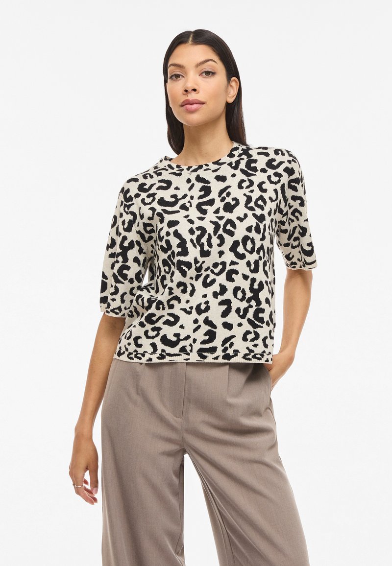 Femme aux longs cheveux noirs portant un haut à manches courtes à imprimé léopard noir et blanc et un pantalon large marron clair, posant avec une main dans la poche.