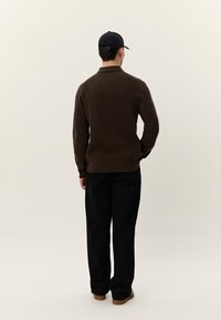 Vyras dėvi tamsiai rudą ribuotą megztinį, juodas kelnes, juodą kepurę ir rudus batus, stovintis viena ranka kišenėje, veidu į kitą pusę.