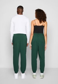 Calças de moletão verdes com punhos elásticos e cintura com cordão, combinadas com uma camisa de manga longa branca e um top preto, vistas por trás.