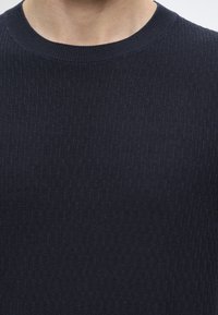 Gros plan d’une personne portant un pull en maille texturée bleu marine foncé à col rond laissant apparaître l’encolure et le haut de la poitrine.
