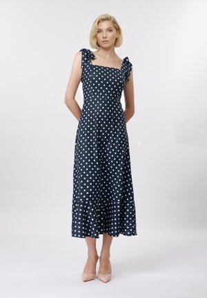 Lyshåret kvinde står iført en marineblå midikjole med hvide polkaprikker, bindebånd på skuldrene og nøgenfarvede spidse hæle mod en ensfarvet baggrund.