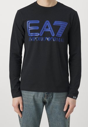 Osoba nosząca czarną bluzkę z długim rękawem z niebieskim logo "EA7 Emporio Armani" i niebieskie dżinsy.
