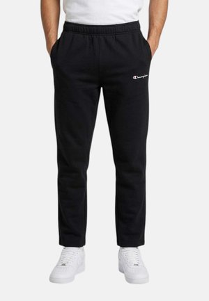 Homme portant un pantalon de survêtement Champion noir avec une taille élastique et des baskets blanches, mains dans les poches, debout devant un fond uni.