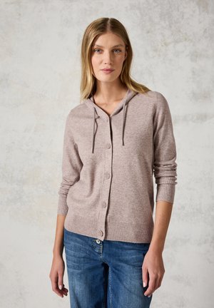 Cardigan rose clair avec une capuche, des poignets côtelés et des boutons à l'avant. Tissu doux et texturé associé à un jean en denim bleu.