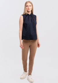 Marineblauw mouwloos topje met geplooide halslijn en sleutelgatdetail, gecombineerd met beige strakke broeken en witte sneakers op een effen achtergrond.