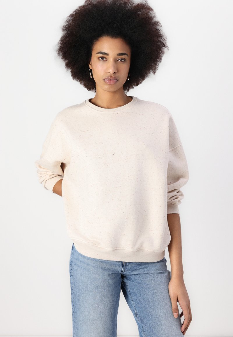 Hellbeige Sweatshirt aus strukturiertem Stoff mit rundem Halsausschnitt und gerippten Bündchen. Kombiniert mit hellblauen Jeans.