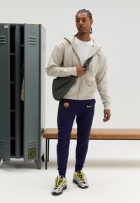 Lichtbeige hoodie met rits, witte T-shirt eronder, donkerpaarse sweatpants met logo, en witte sneakers met zwarte accenten en gele veters.