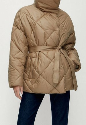 Doudoune matelassée beige avec un col montant et une ceinture assortie nouée à la taille, portée avec un jean bleu foncé.