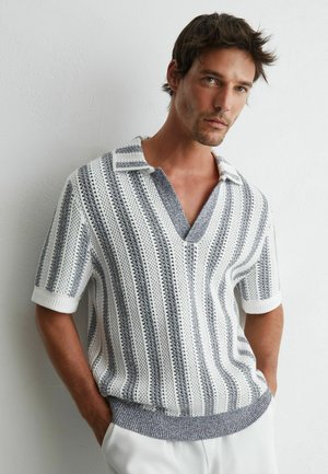 Hombre que lleva una camisa de punto de manga corta a rayas blanca y gris con cuello y escote en V profundo, de pie con las manos en los bolsillos contra una pared blanca.