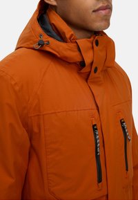 Pomarańczowa ocieplana kurtka z wysokim kołnierzem i regulowanym kapturem, z dwoma przednimi kieszeniami zapinanymi na czarne zamki błyskawiczne oraz kontrastowymi detalami szwów.