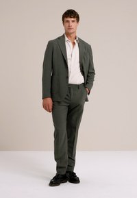 Grau maßgeschneiderter Anzug mit Reverskragen, slim fit Hosen und einem weißen Hemd. Schwarze Schuhe, vor einem neutralen Hintergrund stehend.