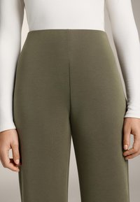 Olívazöld nadrág sima, rugalmas anyagból. Magas derekú kialakítás egyszerű varrással. A modell szűk, fehér hosszú ujjú felsőt visel.