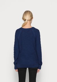 Dunkelblauem Strickpullover mit strukturiertem Muster, Raglanärmeln und breiten Rippbündchen an den Ärmeln und am Saum. Lässige Passform, getragen über schwarzen Hosen.