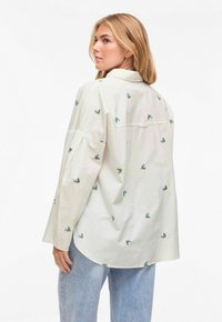 Femme portant une chemise surdimensionnée beige clair avec de petits motifs floraux bleus et un jean bleu clair, regardant par-dessus son épaule gauche.