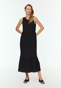 Zwarte mouwloze maxi-jurk met een geraffineerde tiered rok, gemaakt van een lichtgewicht stof. Model gecombineerd met zwarte open-tegen blokhak sandalen.