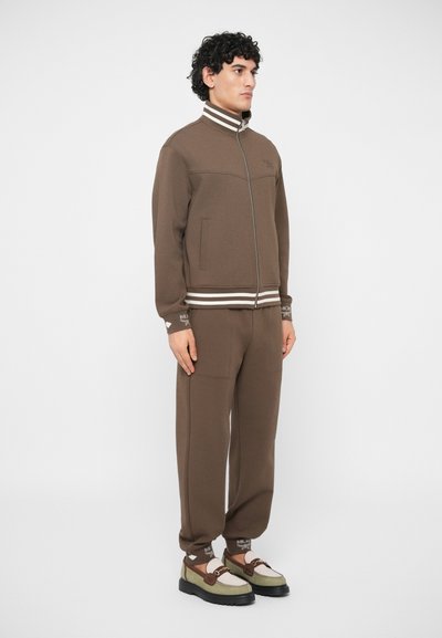 Homem vestindo uma jaqueta castanha com fecho e calças combinando, com gola e punhos listrados, combinados com mocassins verdes e bege.