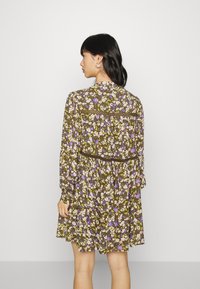 Vestido floral com um fundo verde-azeitona, adornado com flores brancas, roxas e amarelas, mangas longas e uma cintura texturizada e franzida.