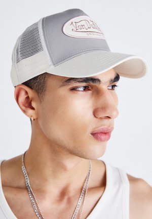 Junger Mann, der eine grau-weiße Netz-Trucker-Cap, ein weißes Tanktop, einen kleinen Creolenohrring und eine silberne Halskette trägt, blickt nach rechts.