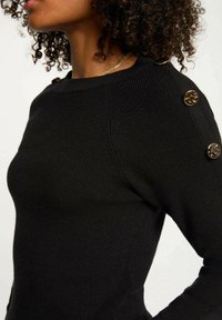 Maglione nero a coste con una vestibilità aderente. Presenta bottoni decorativi dorati sulla spalla e una texture liscia.