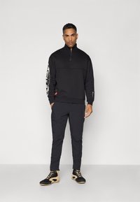 Black half-zip sweatshirt with contrasting white graphic on sleeve, paired with black tapered pants and black, beige-sole athletic shoes.