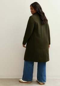 Cappotto in lana verde oliva con taglio dritto, maniche lunghe e spacco posteriore, abbinato a jeans a gamba larga blu e sneakers beige.
