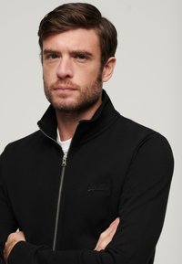 Sort lynlås-sweater lavet af blødt stof med høj krave, der har en diskret logo detalje på brystet og ribbede manchetter.