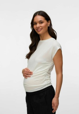 Pregnant woman with long dark hair smiles, wearing a white ruched top and black pants, holding her belly with one hand.