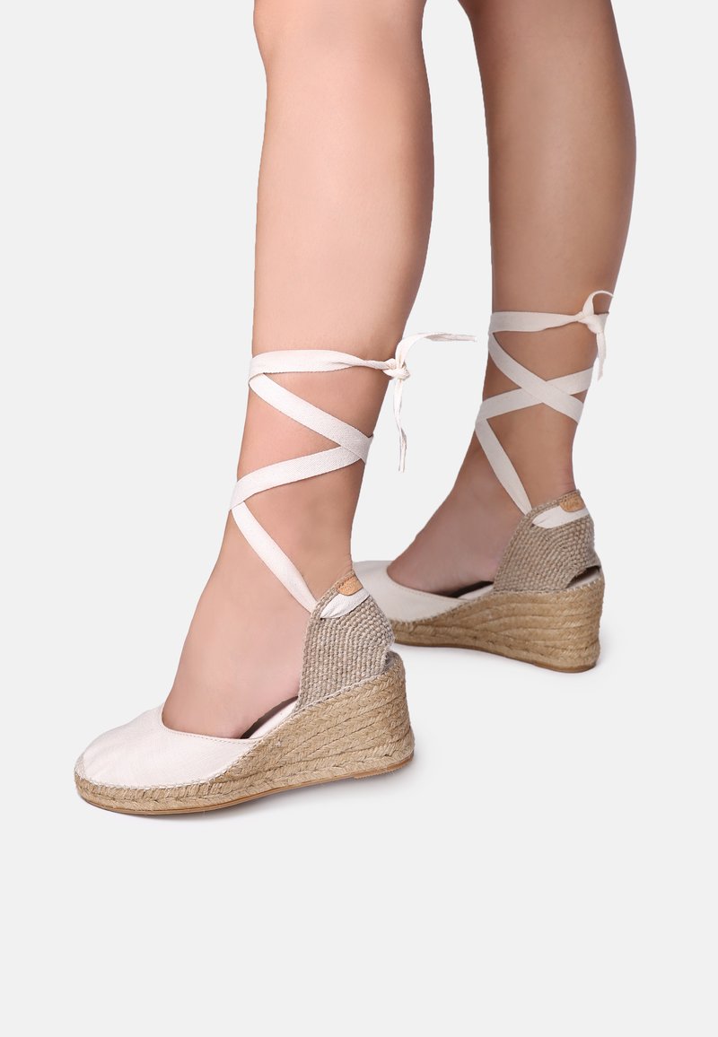 Espadrilles à talons compensés avec un bout arrondi, tige en toile blanche cassée, talon en jute tressé et liens croisées autour de la cheville.
