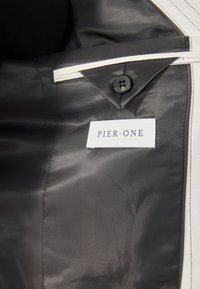 Sort stofinteriør med diagonalt mønster, prydet med en hvid etiket, der står "PIER ONE", og en sort knap.