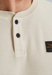 Ecrufarbenes Henley-Shirt aus strukturiertem Stoff, mit drei schwarzen Knöpfen und einem schwarzen Etikett mit goldener Schrift auf der linken Seite.