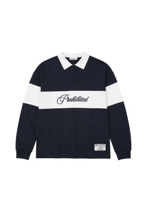 Hombre que lleva una camisa de manga larga en blanco y negro con texto "Prohibited" y una gorra negra que dice "Sunday League Professional", de pie sobre un fondo blanco.