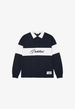 Hombre que lleva una camisa de manga larga en blanco y negro con texto "Prohibited" y una gorra negra que dice "Sunday League Professional", de pie sobre un fondo blanco.