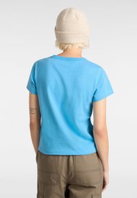 Vaaleansininen lyhythihainen t-paita, jossa pyöreä kaula-aukko; puuvillakangasta. Yhdistetty khaki-värisiin cargo-housuihin; vaalea neulottu pipo. Tekstiyksityiskohta hihassa.