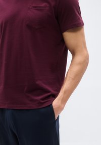 Άτομο φορούν μπορντό μπλουζάκι με κοντά μανίκια και τσέπη στο μπροστινό μέρος και σκούρο μπλε παντελόνι, με το ένα χέρι στην τσέπη, σε μονόχρωμο φόντο.