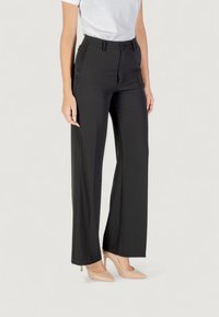 Pantaloni neri a gamba larga con chiusura a bottone e due tasche frontali; realizzati in un tessuto liscio. Indossati con una t-shirt bianca aderente.