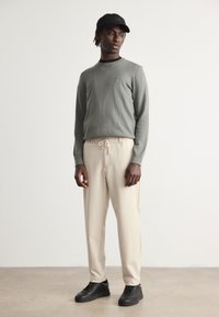 Pull léger gris avec un petit logo sur la poitrine, associé à un pantalon beige avec une taille élastique et des baskets noires. Design minimaliste.