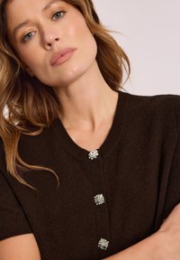 Brązowy dzianinowy kardigan z okrągłym dekoltem, krótkimi rękawami i trzema błyszczącymi guzikami przypominającymi kamienie szlachetne z przodu. Miękka tekstura.