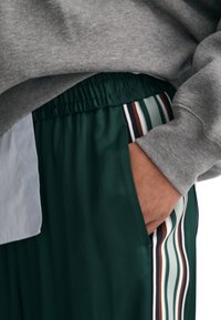 Hand in der Tasche, trägt dunkelgrüne Hosen mit gestreiftem Seitendetail und ein graues langärmliges Hemd, dessen Bündchen über dem Handgelenk sichtbar ist.