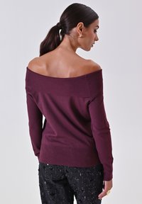 Burgunderfarbener Off-Shoulder-Pullover aus weichem Strickmaterial, mit Rippdetails am Halsausschnitt und figurbetontem Schnitt. Kombiniert mit schwarzen, mit Pailletten besetzten Hosen.