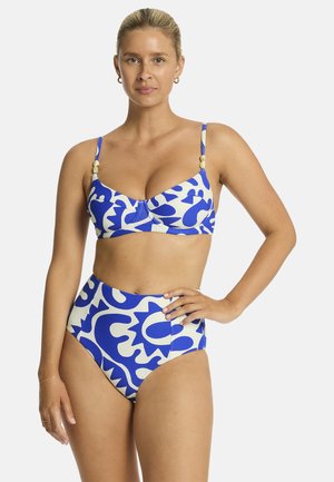Kobieta stojąca z ręką na biodrze, ubrana w niebiesko-biały bikini z abstrakcyjnym wzorem i wysokim stanem, z dekoracyjnymi koralikami na ramiączkach.