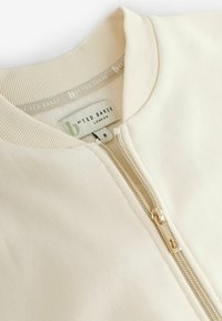 Crèmekleurige ritsjack met ribgebreide kraag en gouden rits, gelabeld "b by Ted Baker London" in maat 8.