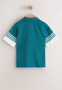 Polo à manches courtes couleur sarcelle avec des poignets blancs présentant trois bandes sarcelles, suspendu sur un cintre en bois contre un mur beige.