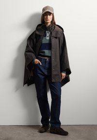 Cape imperméable marron avec un col haut, un pull rayé vert et marine en dessous, un jean en denim foncé, et une casquette à carreaux. Chaussures marron.