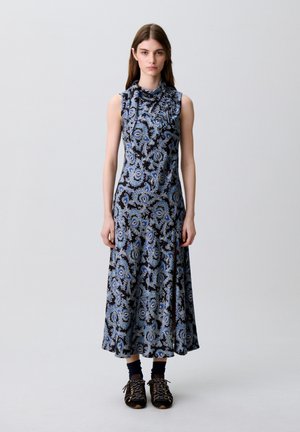 Mujer de pie con un vestido midi sin mangas de patrón cachemir negro y azul, calcetines oscuros y zapatillas negras.