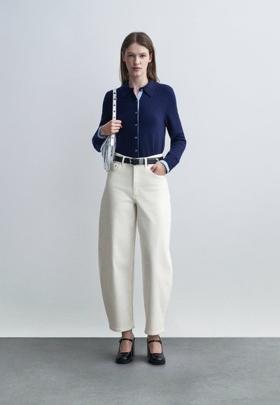 Γυναίκα που στέκεται φορώντας μπλε πουκάμισο με κουμπιά, λευκό παντελόνι φαρδύ σε μέση υψηλή, μαύρες γόβες και κρατώντας ασημένια τσάντα στον ώμο.